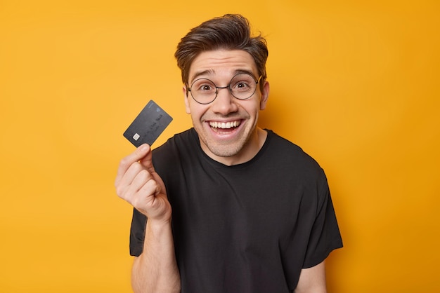 Financial services concept Positive brunet adult man holds credit card uses electronic money happy to get money on account wears round spectcles and black t shirt isoated over yellow background