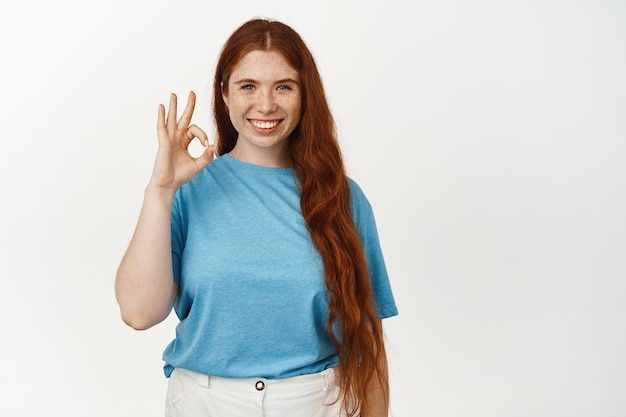 Fine, not bad. Smiling redhead female showing okay OK sign, say yes, nod in approval, agree with you, praise good work, like and agree with excellent choice, white background.