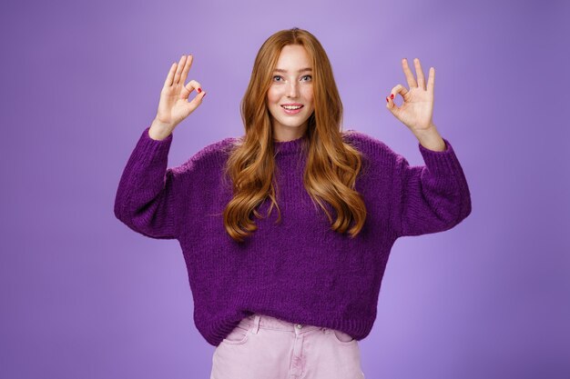 Fine I agree. Portrait of friendly and optimstic young 20s ginger girl in purple sweater raising hands with okay or ok gesture smiling in approval, liking cool product, giving recommendation.