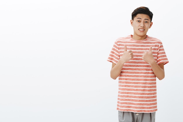 Fine I guess. Portrait of unsure awkward young attractive asian man in striped t-shirt making tight ucertain smile and showing thumbs up gesture as if agree or like idea