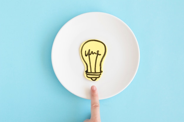 Finger pointing toward paper cutout light bulb on white dish against blue background