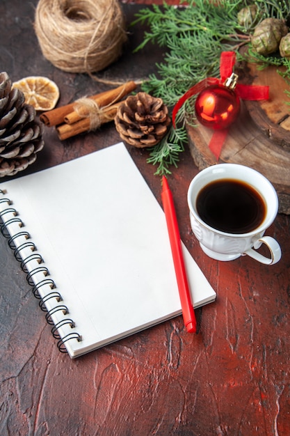 Fir branches and closed spiral notebook with pen cinnamon limes conifer cone and a cup of black tea on dark background