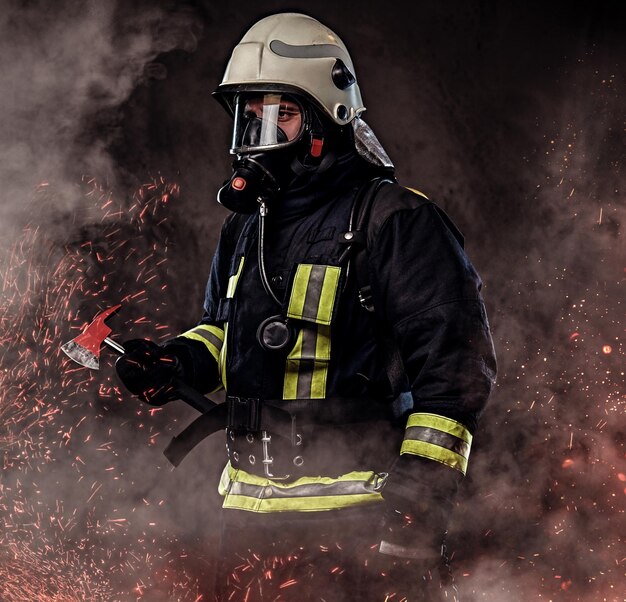 A firefighter dressed in a uniform and an oxygen mask holds a red axe standing in fire sparks and smoke over a dark background.