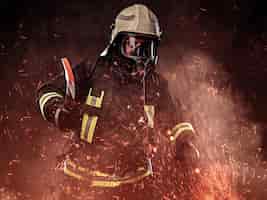 Foto gratuita un vigile del fuoco vestito con un'uniforme e una maschera di ossigeno tiene un'ascia rossa in piedi in scintille di fuoco e fumo su uno sfondo scuro.