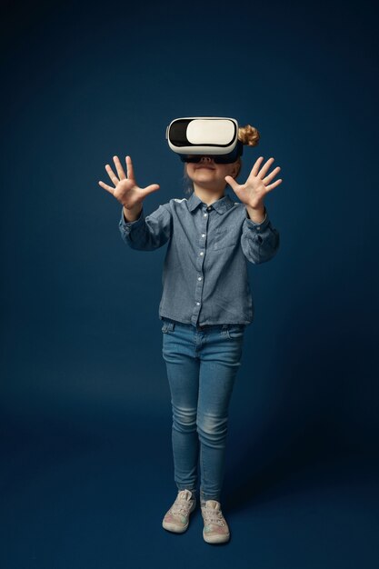 First steps in fairy. Little girl or child in jeans and shirt with virtual reality headset glasses isolated on blue studio background. Concept of cutting edge technology, video games, innovation.