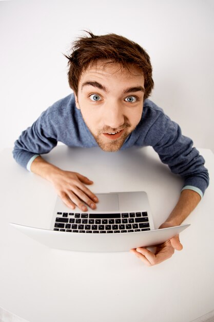 fish-eye upper angle shot of funny guy, staring with smile, using laptop, working on project, typing on computer