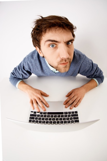 Fish-eye upper angle shot of thoughtful, suspicious young man having doubts, staring with disbelief, working with laptop