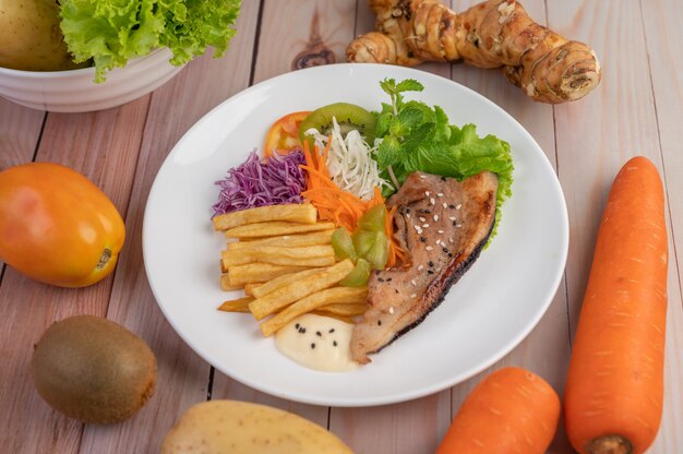 Fish steak with french fries, kiwi, lettuce, carrots, tomatoes, and cabbage in a white dish.