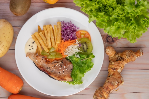Стейк из рыбы с картофелем фри, киви, листьями салата, морковью, помидорами и капустой в белом блюде.