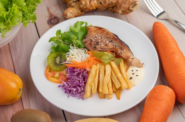 Fish steak with french fries, kiwi, lettuce, carrots, tomatoes, and cabbage in a white dish.