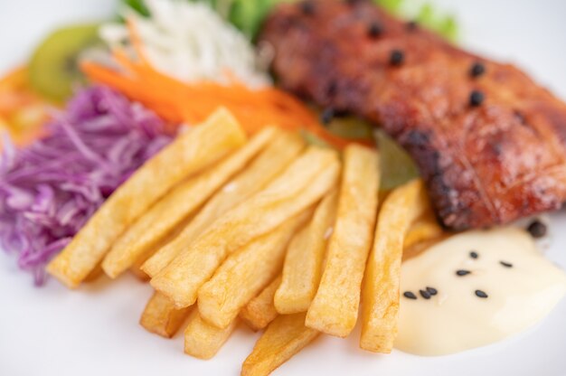 Fish steak with french fries, kiwi, lettuce, carrots, tomatoes, and cabbage in a white dish.