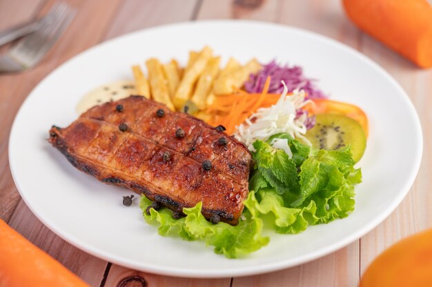 Fish steak with french fries, kiwi, lettuce, carrots, tomatoes, and cabbage in a white dish.