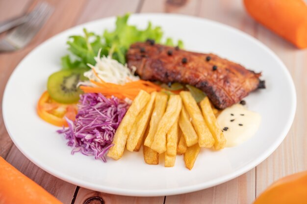 Fish steak with french fries, kiwi, lettuce, carrots, tomatoes, and cabbage in a white dish.