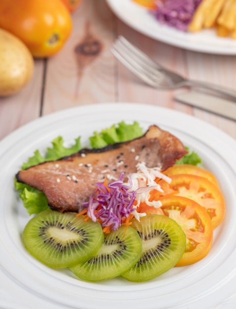 Fish steak with french fries, kiwi, lettuce, carrots, tomatoes, and cabbage in a white dish.