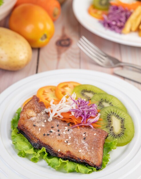 Fish steak with french fries, kiwi, lettuce, carrots, tomatoes, and cabbage in a white dish.