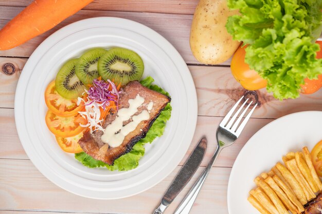 Fish steak with french fries, kiwi, lettuce, carrots, tomatoes, and cabbage in a white dish.