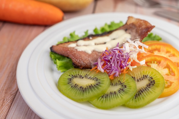 Fish steak with french fries, kiwi, lettuce, carrots, tomatoes, and cabbage in a white dish.