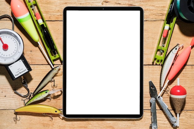 Fishing lure; fishing float; plier and weighing scale near the digital tablet with white screen on desk