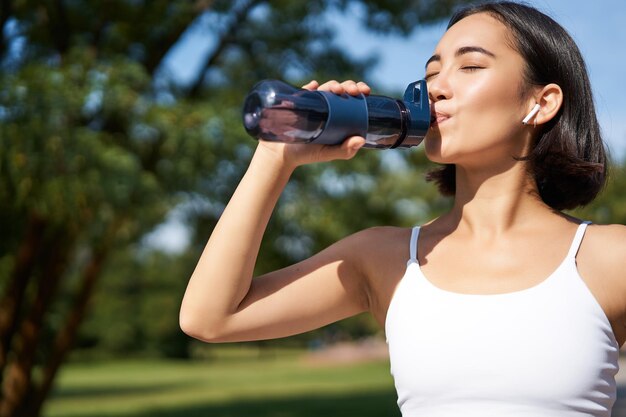 Fit asian sportswoman drinks water while running marathon fitness girl jogging in park staying hydra