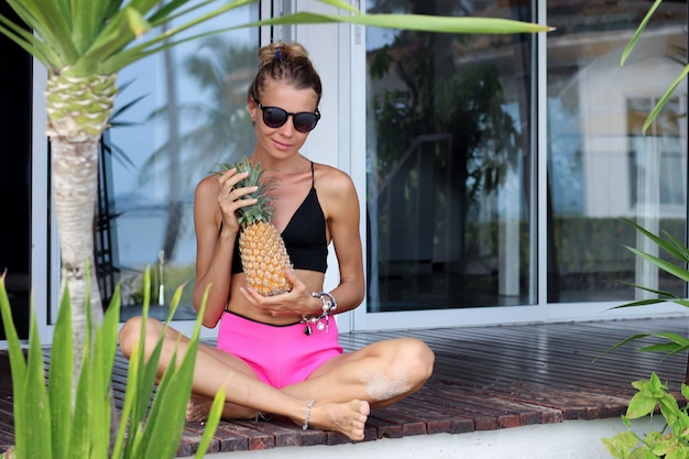 Fit caucasian woman in black top pink shorts hold pineapple outside tropical villa