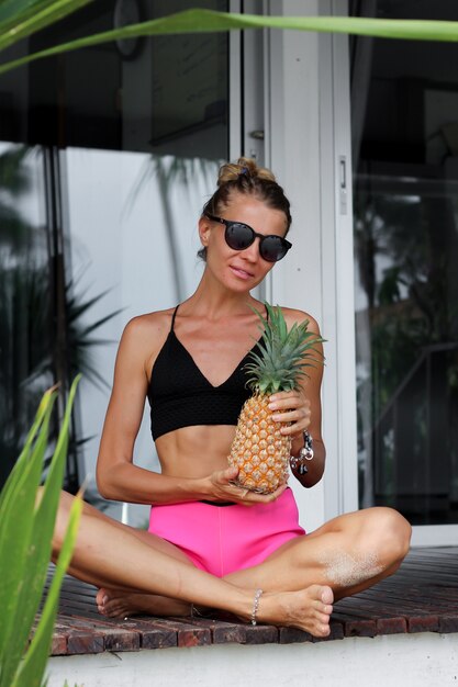 Fit caucasian woman in black top pink shorts hold pineapple outside tropical villa