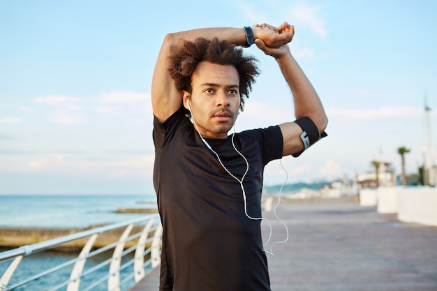 Fit dark-skinned male athlete with bushy hair doing stretching exercises, raising his arms, warming up his muscles before outdoor morning workout session