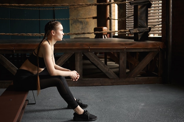 Fit girl having rest after intensive cardio workout in gym. Sideways portrait of tired serious young female boxer in black running shoes and sports outfit relaxing on bench