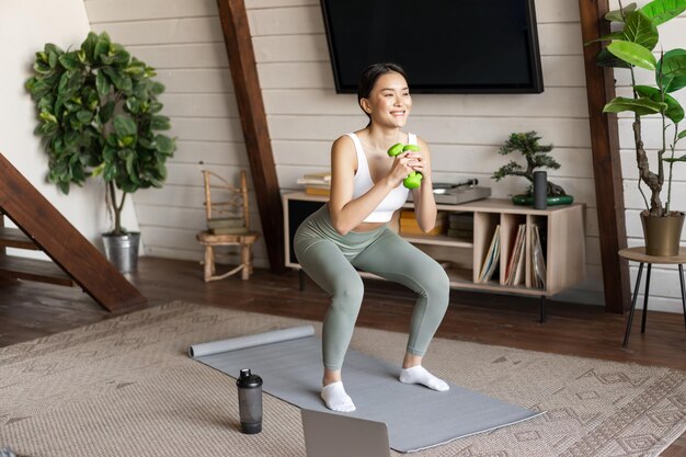 Fit and healthy asian woman doing squats at home workout fitness training session with dumbbells and...