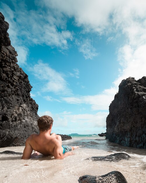 Fit male laying and relaxing on the sandy beach near large black rocks and looking at the sea