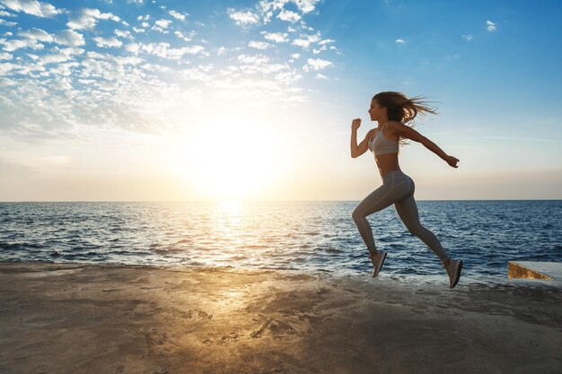 Fit sporty stylish caucasian young sportswoman fitness instructor leading active lifestyle jumping making cardio exercise near seaside docks enjoying sunset workout wearing sportswear