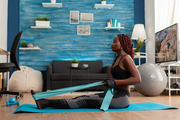 Подходит для сильной афро-женщины, тренирующейся в домашней гостиной, используя эспандер, сидя на коврике для фитнеса, потянув за мышцы спины
