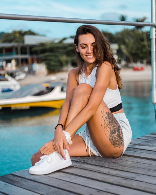 Fit tanned brunette tattooed woman in light blue jean shorts and white fitting top sits on wooden pier at sunset light
