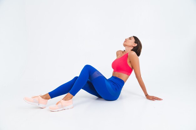 Fit tanned sporty woman with abs, fitness curves, wearing top and blue leggings on white