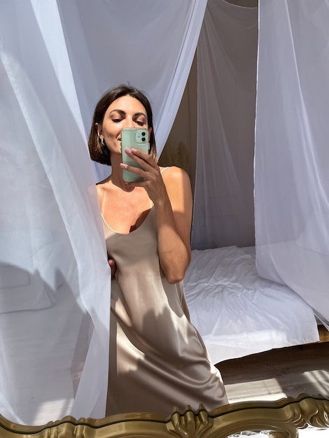 Free Photo fit tanned woman in romantic beige silk dress at home takes photo selfie on phone in mirror