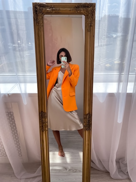 Una donna abbronzata in forma in un romantico abito di seta beige e un blazer arancione a casa scatta una foto selfie al telefono allo specchio