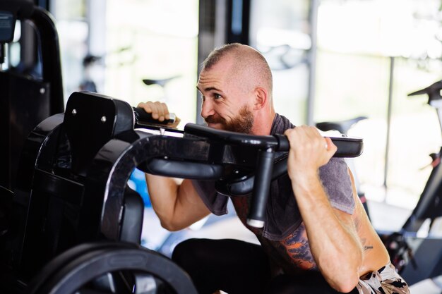 Fit tattooed bearded man doing squats in a training machine