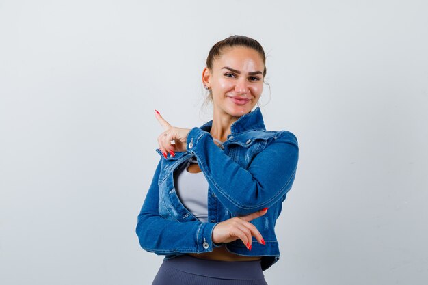 Fit woman in crop top, jean jacket, leggings pointing left and right with index fingers and looking cheery , front view.