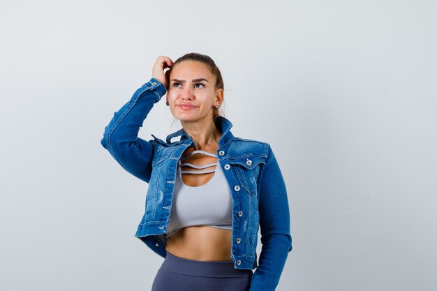 Fit woman in crop top, jean jacket, leggings scratching head, thinking about something and looking pensive , front view.