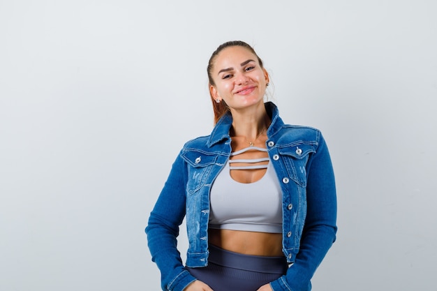 Fit woman in crop top, jean jacket, leggings smiling while posing at camera and looking cheerful , front view.