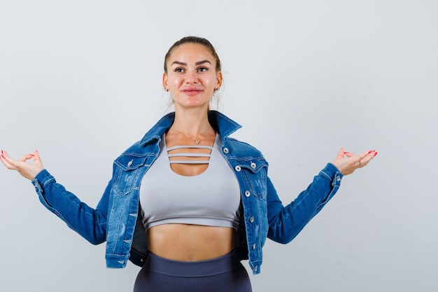 Fit woman in crop top, jean jacket, leggings standing in meditating pose and looking cheery , front view.