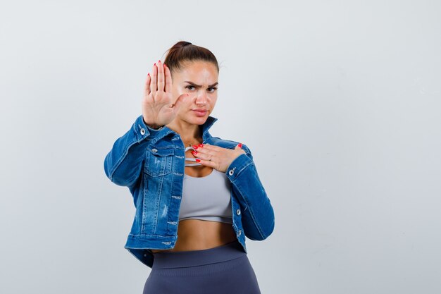Fit woman showing stop sign, with hand over chest in crop top, jean jacket, leggings and looking confident , front view.