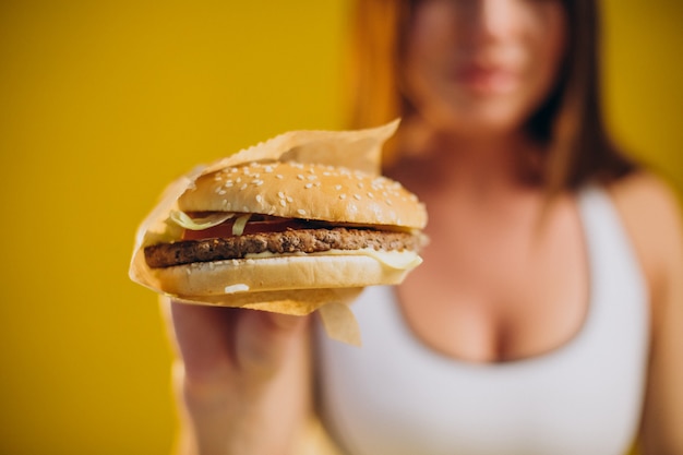 Подходящая женщина в спортивной одежде ест гамбургер, изолированные на желтом фоне