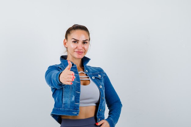 Fit woman stretching hand toward camera to greet, with hand on hip in crop top, jean jacket, leggings and looking amiable. front view.