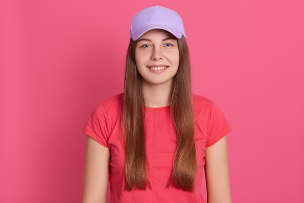 バラ色の壁の上に孤立した立っているカジュアルなtシャツを着ている若い女性に合います。野球帽の美しいモデル