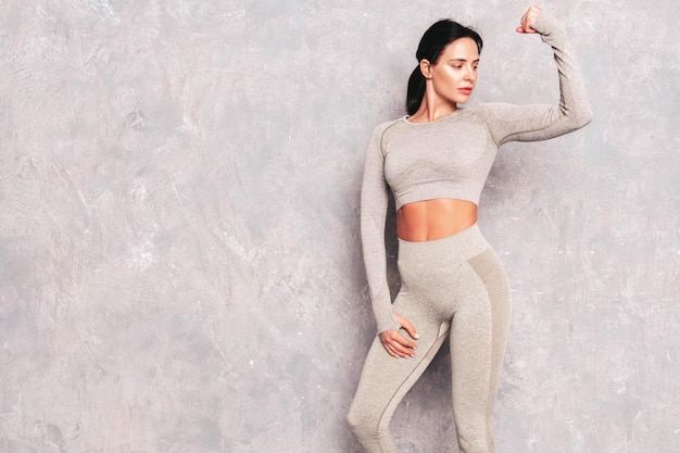 Fitness confident woman in sports clothing Sexy young beautiful model with perfect body Female posing near grey wall in studio Stretching out before trainingShows her muscles