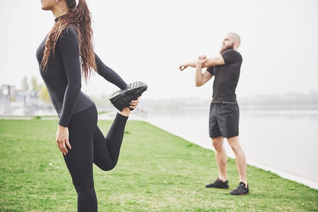 水の近くの公園で屋外ストレッチフィットネスカップル。若いあごひげを生やした男性と女性が朝一緒に運動