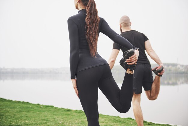 水の近くの公園で屋外ストレッチフィットネスカップル。若いあごひげを生やした男性と女性が朝一緒に運動