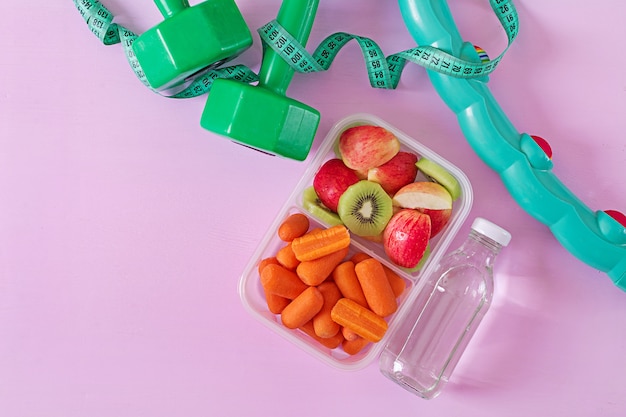 Fitness equipment. Healthy food. Concept healthy food and sports lifestyle. Vegetarian lunch. Dumbbell, water, fruits on pink surface. Top view. Flat lay