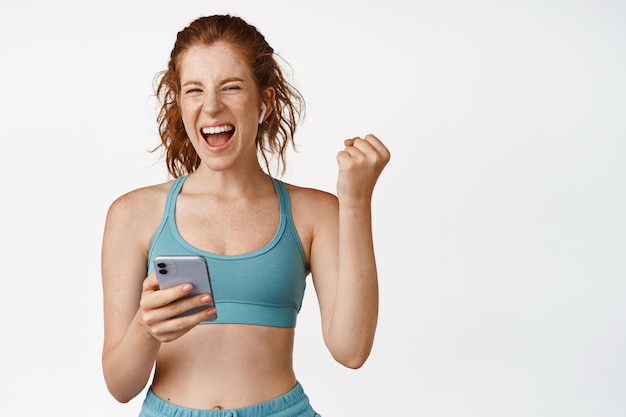 Foto gratuita la ragazza di forma fisica raggiunge l'obiettivo dell'app per lo sport sul telefono cellulare urlando soddisfatto e sorridente soddisfatto allenamento in abbigliamento sportivo in piedi su sfondo bianco