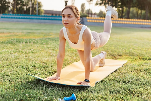 屋外スタジアムでヨガマットの脚のトレーニングをしているフィットネスの女の子、白いトップとベージュのレギンスを一人でトレーニングしているフィットの女性、ヘルスケア、健康的なライフスタイル。